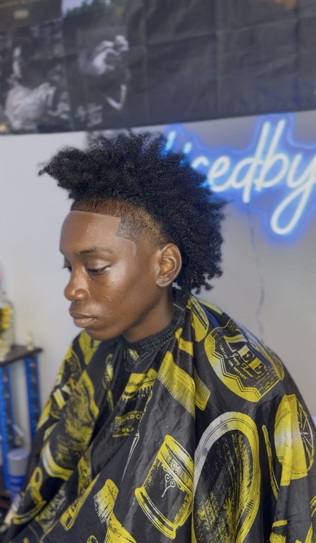 Youth client with a high-top afro, sharp forehead lineup and temple fade in front of a Sliced by Re neon sign